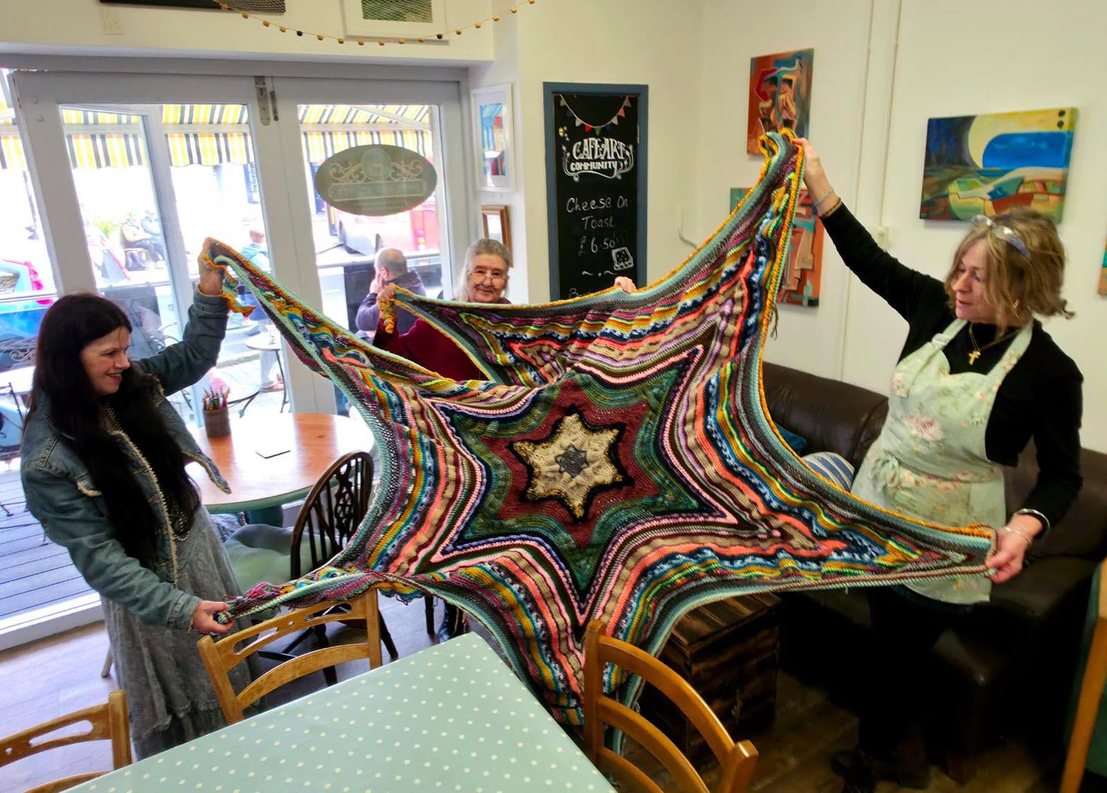 Cafe Art regulars holding a large colourful crocheted community artwork